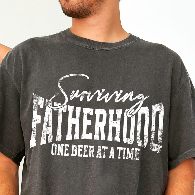 Puede incluir: Una camiseta gris oscura con un gr&aacute;fico blanco desgastado que dice "Surviving Fatherhood One Beer at a Time."