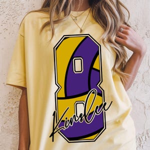 May include: Yellow t-shirt featuring a large number 8 in yellow and purple, with the name "Kinslee" in a cursive font. The design is outlined in black. The shirt is a light yellow color.