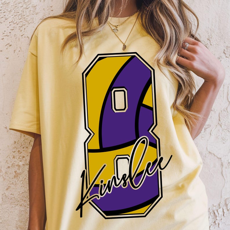 May include: Yellow t-shirt featuring a large number 8 in yellow and purple, with the name "Kinslee" in a cursive font. The design is outlined in black. The shirt is a light yellow color.