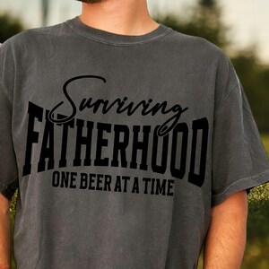 Puede incluir: Una camiseta gris con texto negro que dice "Surviving Fatherhood One Beer at a Time".