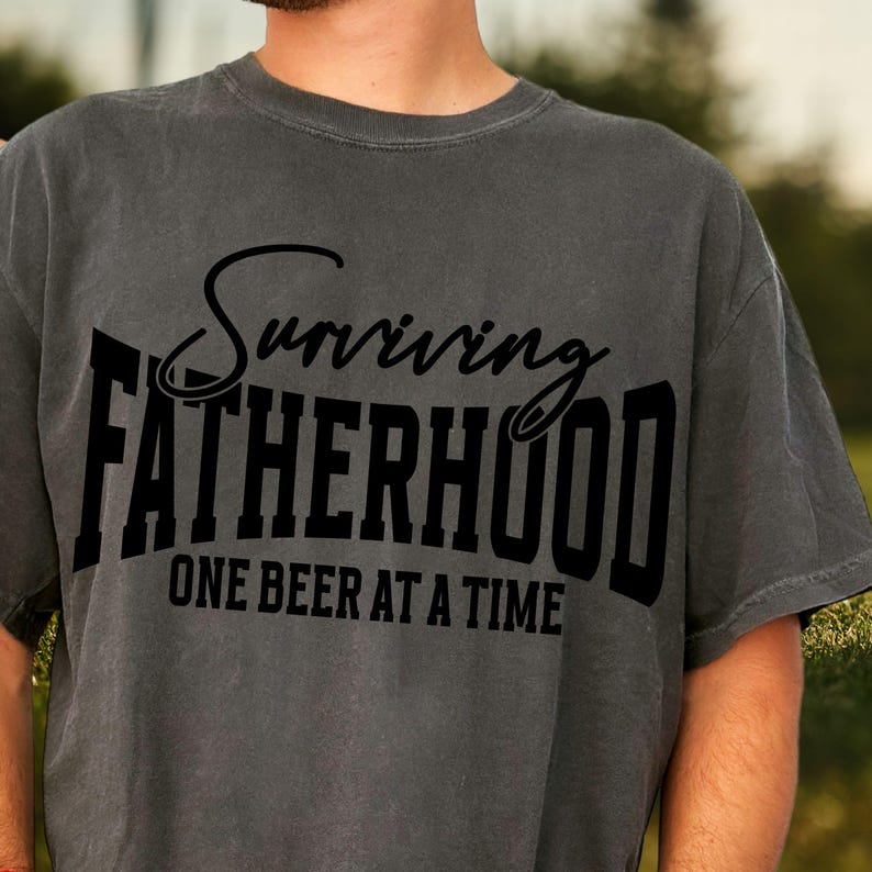 Puede incluir: Una camiseta gris con texto negro que dice "Surviving Fatherhood One Beer at a Time".