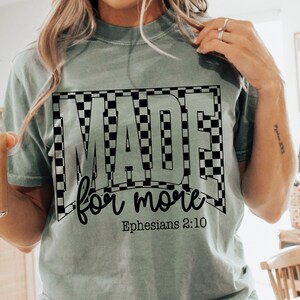 May include: A light green t-shirt with the words "MADE for more" in a black and white checkered design. The text "Ephesians 2:10" is below the main text. The shirt has short sleeves.