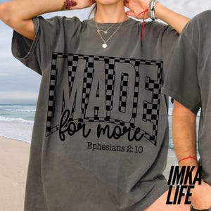 May include: Dark gray t-shirt with the words "MADE for more" in a black checkered design. The text "Ephesians 2:10" is below the main text. The shirt is a comfortable, casual style.
