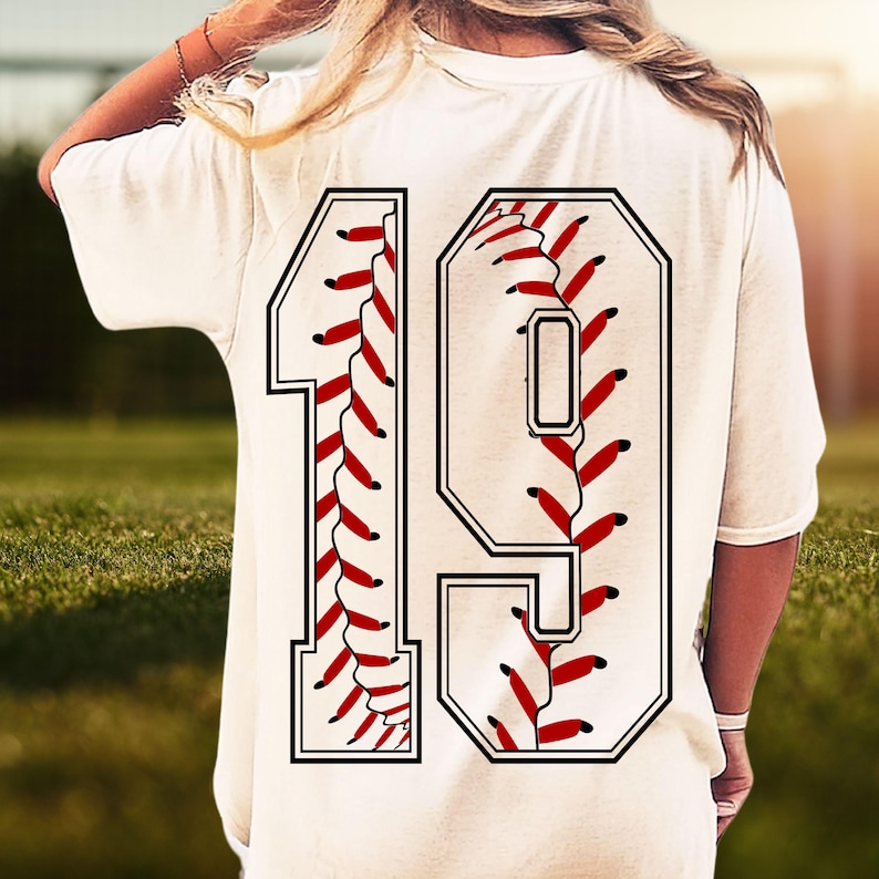 May include: Cream-colored t-shirt with a large baseball-themed number 19 on the back. The number is white with a black outline and red stitching details, resembling a baseball. The shirt is worn outdoors.