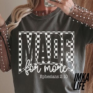 May include: Dark gray t-shirt with the words "MADE for more" in a white, checkered design. The text "Ephesians 2:10" is below the main text. The shirt also has the text "IMKA LIFE" in the bottom right corner.