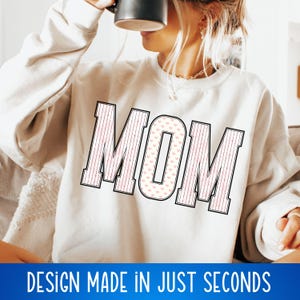 May include: A white sweatshirt with the word "MOM" printed in black outlined letters. The letters are filled with a pink and white heart pattern.
