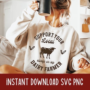 Puede incluir: Una sudadera blanca con el texto "Support Your Local Dairy Farmer" en letras marrones. Una silueta de vaca marrón se encuentra en el centro del texto. El texto "always on tap" se encuentra debajo de la vaca.