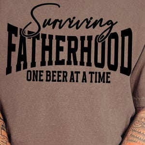 Puede incluir: Una camiseta marr&oacute;n con texto negro que dice "Surviving Fatherhood One Beer at a Time".
