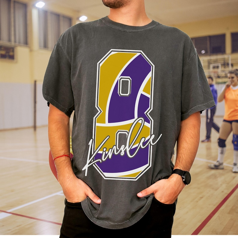 May include: Dark gray t-shirt featuring a large number 8 graphic in gold and purple, with the name "Kinslee" in a cursive font. The shirt is worn by a person with their hands in their pockets, with a volleyball court in the background.