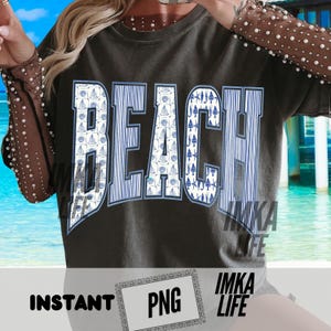 Op de afbeelding: Donkergrijs t-shirt met het woord "BEACH" in grote, vette letters. De letters zijn gevuld met een patroon van blauwe en witte zeilboten en verticale strepen. Het shirt heeft ook de tekst "IMKA LIFE" en "INSTANT PNG".