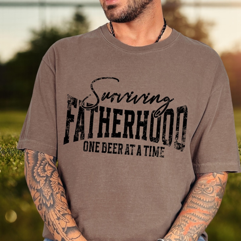 Puede incluir: Una camiseta marr&oacute;n con un gr&aacute;fico negro desgastado que dice "Surviving Fatherhood One Beer at a Time".