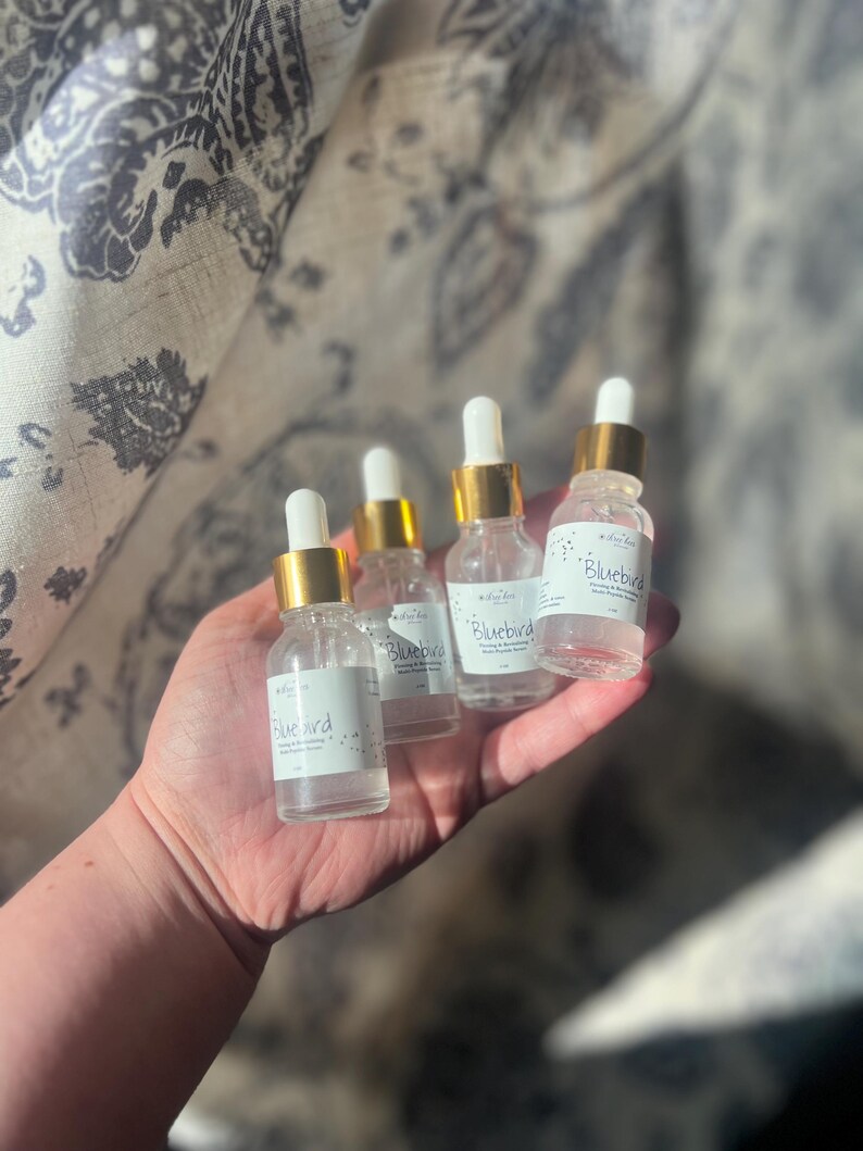K&ouml;nnte beinhalten: Vier kleine Glasflaschen mit goldenen Kappen und Etiketten mit der Aufschrift "Bluebird", die eine klare Fl&uuml;ssigkeit enthalten. Die Flaschen werden in der Hand einer Person gehalten.
