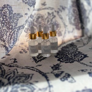 K&ouml;nnte beinhalten: Vier kleine Glasflaschen mit goldenen Kappen und klarer Fl&uuml;ssigkeit im Inneren. Die Flaschen sind mit "Bluebird" beschriftet und stehen auf einem wei&szlig;en Stoff mit blauem Blumenmuster.