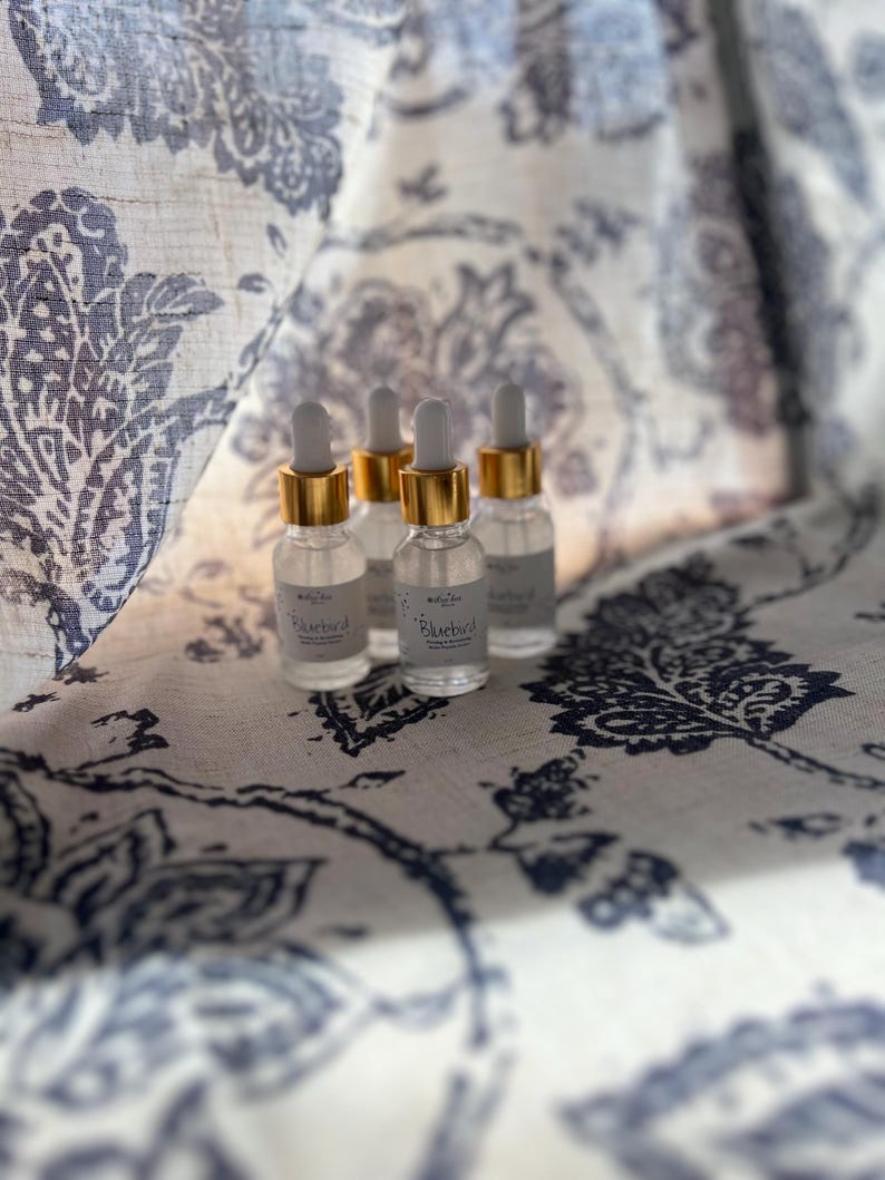 K&ouml;nnte beinhalten: Vier kleine Glasflaschen mit goldenen Kappen und klarer Fl&uuml;ssigkeit im Inneren. Die Flaschen sind mit "Bluebird" beschriftet und stehen auf einem wei&szlig;en Stoff mit blauem Blumenmuster.