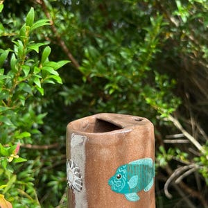 May include: A brown ceramic cup with a turquoise fish design and a sunburst pattern. The cup has a rectangular shape with a curved top and is held against a backdrop of green foliage.
