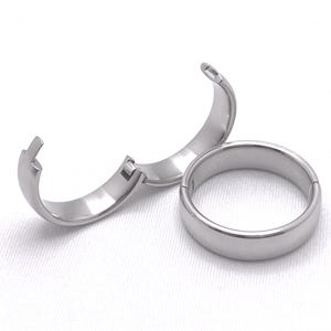 May include: Two silver rings, one with a hinged opening, are shown on a white surface. The rings are both plain and smooth.
