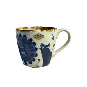 May include: A white ceramic mug with a blue and brown floral design and a black cat illustration. The mug has a brown rim and a handle. The text "有田焼 ARITA" is printed on the mug.