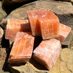 May include: Orange calcite crystal chunks. The raw, natural stones have a translucent, crystalline structure. The pieces vary in size and shape, with a warm, earthy color. The crystals are displayed on a bed of rocks and wood.