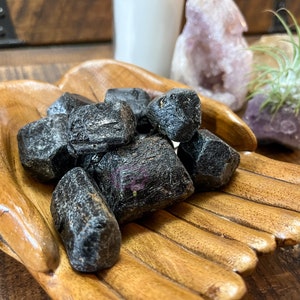 May include: A collection of black tourmaline crystals in a wooden hand-shaped bowl. The crystals are rough and have a natural, unpolished look.