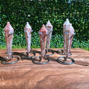 May include: Six pointed, multi-faceted stone pendulums with swirling patterns of cream, tan, and reddish-brown. Each pendulum is secured in a circular silver-toned metal wire stand, displaying a polished, smooth texture and a tapered, crystalline shape.