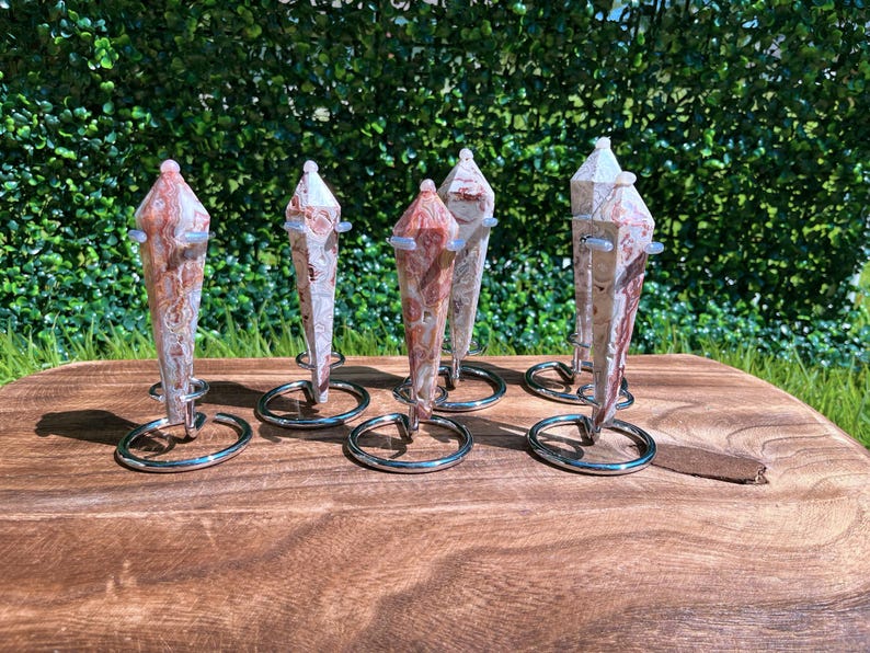 May include: Six pointed, multi-faceted stone pendulums with swirling patterns of cream, tan, and reddish-brown. Each pendulum is secured in a circular silver-toned metal wire stand, displaying a polished, smooth texture and a tapered, crystalline shape.