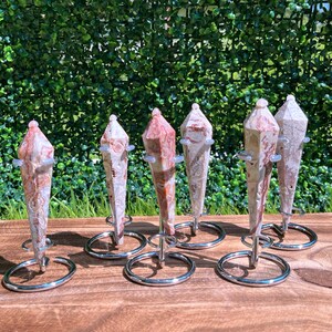 May include: Six pointed, faceted stone pendulums with marbled patterns in shades of cream, tan, and reddish-brown. Each pendulum is suspended vertically within a circular, silver-toned metal wire stand, showcasing a polished, smooth surface texture.