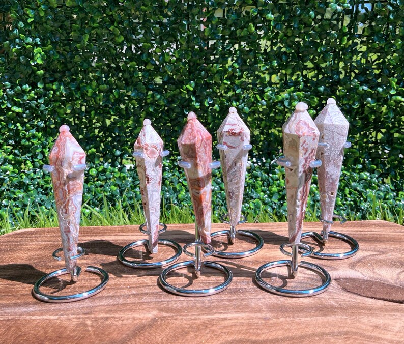 May include: Six pointed, faceted stone pendulums with marbled patterns in shades of cream, tan, and reddish-brown. Each pendulum is suspended vertically within a circular, silver-toned metal wire stand, showcasing a polished, smooth surface texture.