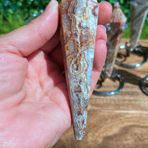 May include: A faceted, pointed crystal pendulum featuring intricate, swirling bands of brown, white, and translucent gray. The stone has a polished, smooth surface with a distinct layered pattern throughout its elongated, tapered, and symmetrical shape.