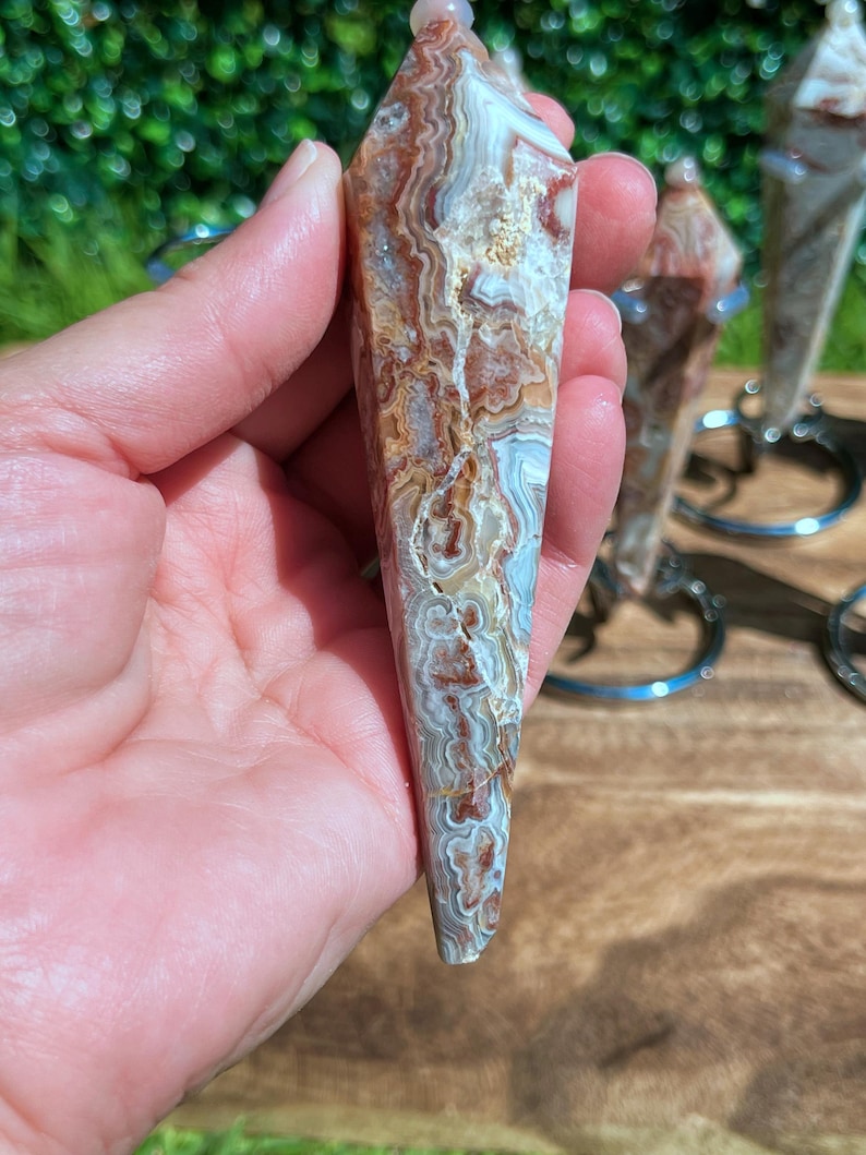 May include: A faceted, pointed crystal pendulum featuring intricate, swirling bands of brown, white, and translucent gray. The stone has a polished, smooth surface with a distinct layered pattern throughout its elongated, tapered, and symmetrical shape.