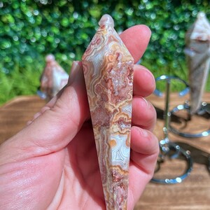 May include: A faceted, pointed crystal pendulum made of banded agate with intricate, swirling patterns in shades of reddish-brown, cream, and white. The stone has a polished, smooth surface and a tapered, hexagonal shape that comes to a sharp point.