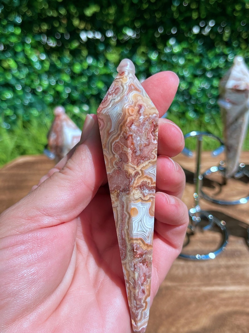 May include: A faceted, pointed crystal pendulum made of banded agate with intricate, swirling patterns in shades of reddish-brown, cream, and white. The stone has a polished, smooth surface and a tapered, hexagonal shape that comes to a sharp point.