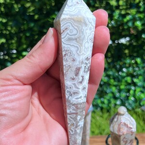 May include: A polished, hexagonal-shaped crystal pendulum featuring intricate, swirling patterns in shades of cream, beige, and soft brown. The stone has a smooth, glossy finish and tapers to a point at the bottom, topped with a small, rounded bead.
