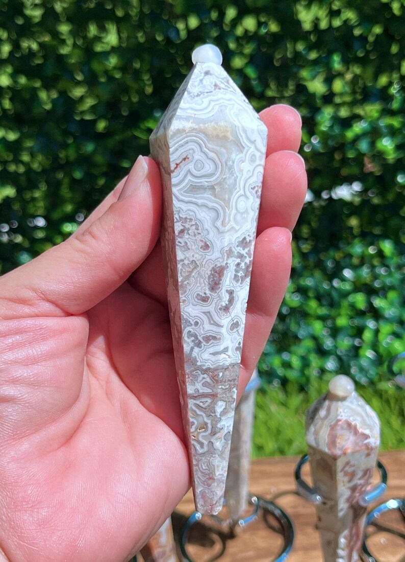 May include: A polished, hexagonal-shaped crystal pendulum featuring intricate, swirling patterns in shades of cream, beige, and soft brown. The stone has a smooth, glossy finish and tapers to a point at the bottom, topped with a small, rounded bead.