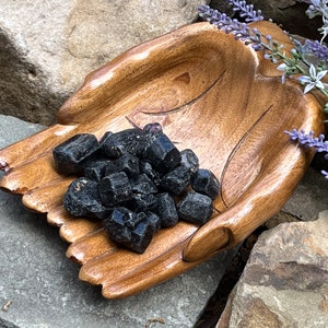 May include: A wooden hand-shaped bowl filled with black tourmaline crystals. The bowl is made of dark wood and has a natural finish. The crystals are rough and have a black color.