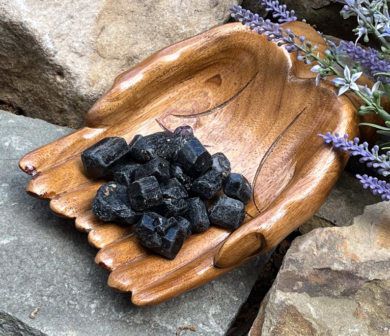 May include: A wooden hand-shaped bowl filled with black tourmaline crystals. The bowl is made of dark wood and has a natural finish. The crystals are rough and have a black color.