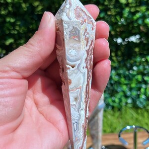 May include: A polished, multi-faceted crystal tower featuring intricate, swirling bands of white, cream, tan, and reddish-brown agate. The stone has a pointed top and a smooth, elongated hexagonal shape with visible natural mineral inclusions.