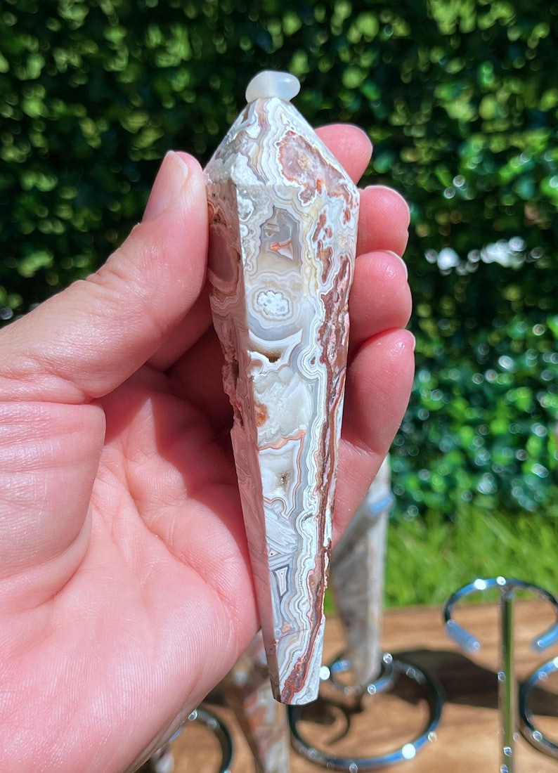 May include: A polished, multi-faceted crystal tower featuring intricate, swirling bands of white, cream, tan, and reddish-brown agate. The stone has a pointed top and a smooth, elongated hexagonal shape with visible natural mineral inclusions.