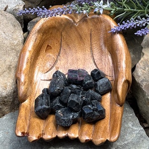 May include: A wooden hand-shaped bowl filled with black tourmaline crystals. The bowl is made of dark wood and has a natural finish. The crystals are a deep black color and have a shiny, almost metallic finish.