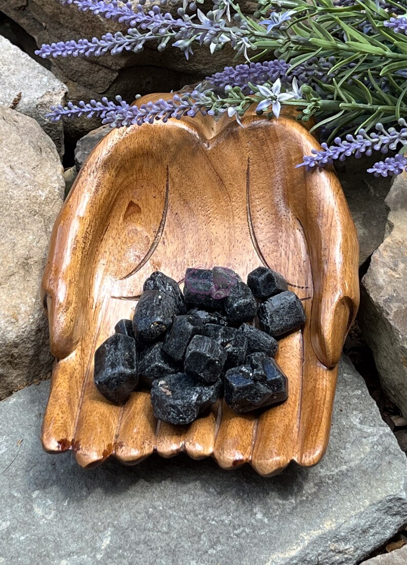 May include: A wooden hand-shaped bowl filled with black tourmaline crystals. The bowl is made of dark wood and has a natural finish. The crystals are a deep black color and have a shiny, almost metallic finish.