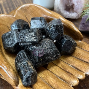 May include: A collection of black, rough-cut, natural crystals in a wooden bowl. The crystals are arranged in a pile, with some overlapping.