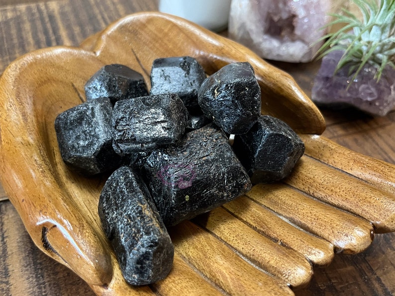 May include: A collection of black, rough-cut, natural crystals in a wooden bowl. The crystals are arranged in a pile, with some overlapping.