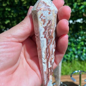May include: A faceted, pointed crystal pendulum featuring intricate, swirling patterns of cream, beige, and deep reddish-brown. The stone has a polished, smooth surface with a distinct banded texture and a small, rounded white bead at the top point.