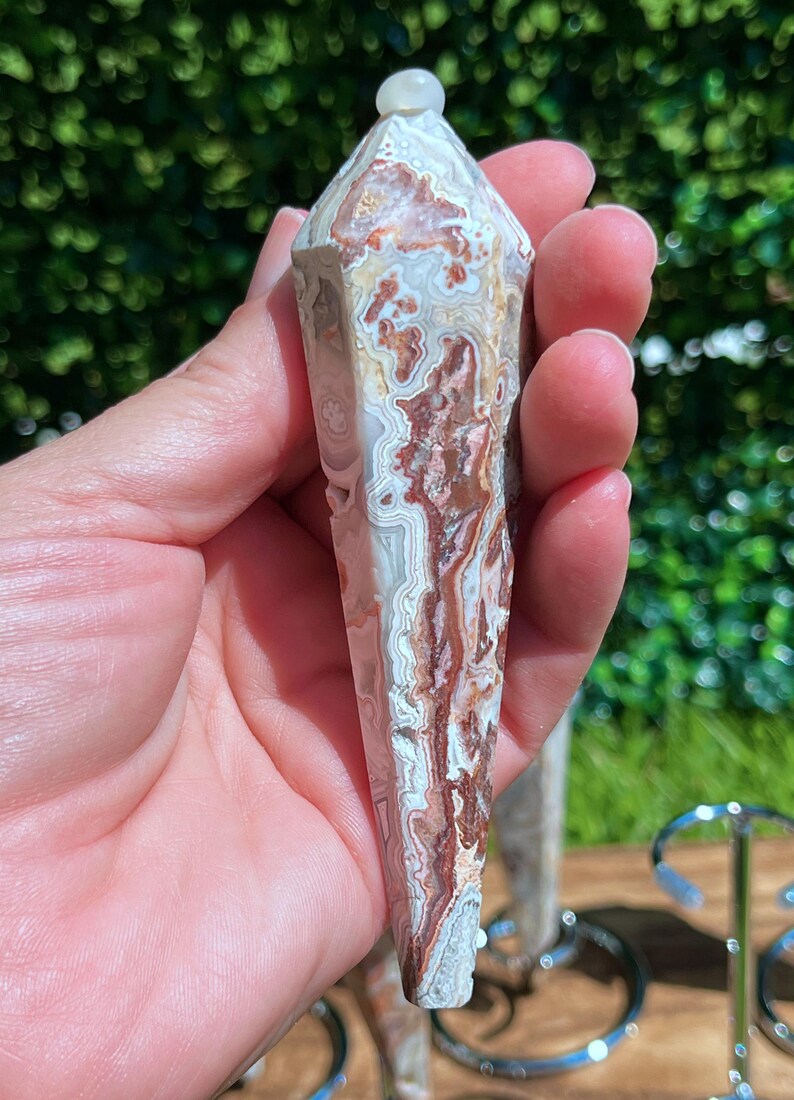 May include: A faceted, pointed crystal pendulum featuring intricate, swirling patterns of cream, beige, and deep reddish-brown. The stone has a polished, smooth surface with a distinct banded texture and a small, rounded white bead at the top point.