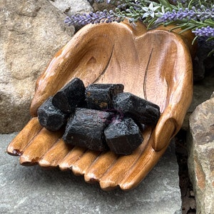 May include: A wooden hand-shaped bowl filled with black tourmaline crystals. The bowl is made of dark wood and has a smooth, polished finish. The crystals are rough and have a natural, unpolished look.
