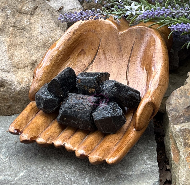 May include: A wooden hand-shaped bowl filled with black tourmaline crystals. The bowl is made of dark wood and has a smooth, polished finish. The crystals are rough and have a natural, unpolished look.