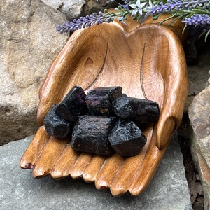 May include: A collection of black obsidian stones in a wooden hand-shaped bowl. The bowl is resting on a gray rock.