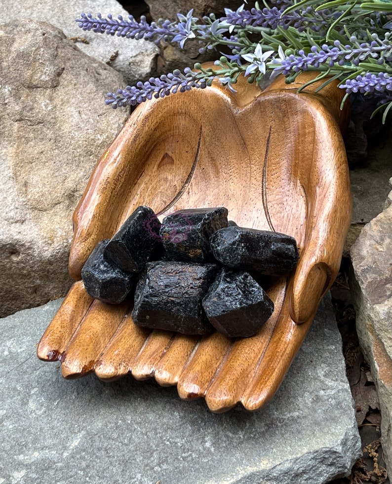 May include: A collection of black obsidian stones in a wooden hand-shaped bowl. The bowl is resting on a gray rock.
