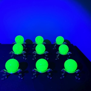 Puede incluir: Siete esferas de vidrio verde brillante sobre soportes metálicos ornamentados. Las esferas están dispuestas en un patrón de cuadrícula sobre una superficie oscura, iluminadas por luz azul. Los soportes tienen un diseño decorativo, posiblemente antiguo.