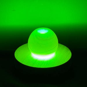 May include: A glowing green orb sits atop a matching circular base, illuminated with a bright white light. The sphere has a textured surface, and the background is a gradient of green.
