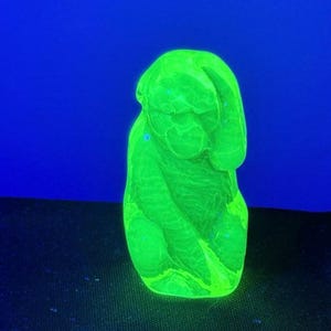 May include: A glowing green glass sculpture of a figure, possibly a torso or abstract form, against a blue background. The glass has a textured surface and appears to be illuminated from within, creating a vibrant, eye-catching effect.